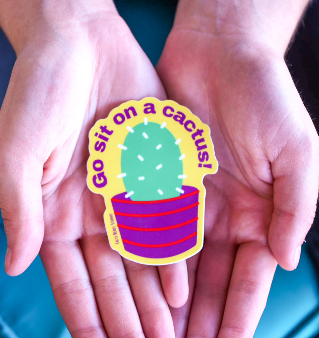 Colorful sticker with a cactus and text held between two hands against a blue background